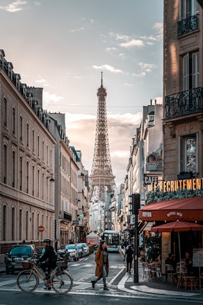 A bustling Parisian street scene with the Eiffel Tower visible in the distance. People are walking and cycling along the street lined with classic European buildings. A caf&eacute; with a red canopy and pedestrians crossing the road add to the lively atmosphere.