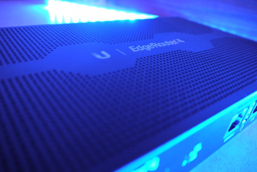A close-up view of a network device with a perforated surface, labeled 'EdgeRouter 4' under blue LED lighting. The image shows parts of the device's ports and connections on the side.
