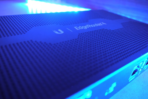 A close-up view of a network device with a perforated surface, labeled 'EdgeRouter 4' under blue LED lighting. The image shows parts of the device's ports and connections on the side.