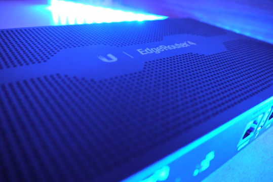 A close-up view of a network device with a perforated surface, labeled 'EdgeRouter 4' under blue LED lighting. The image shows parts of the device's ports and connections on the side.