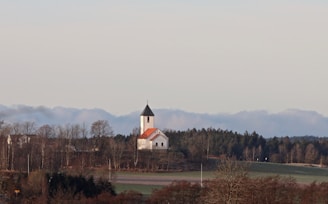 A small church with a red roof and white walls stands atop a gentle hill, surrounded by bare trees and patches of green grass. The sky is overcast, and there are low-lying clouds in the distance, adding a sense of calm and serenity to the scene.