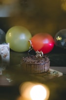 A chocolate cake adorned with a number '30' candle sits on a wooden board. Surrounding it are balloons in various colors including yellow, red, and transparent. A greeting card is partially visible on the left side.