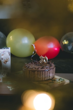 A chocolate cake adorned with a number '30' candle sits on a wooden board. Surrounding it are balloons in various colors including yellow, red, and transparent. A greeting card is partially visible on the left side.