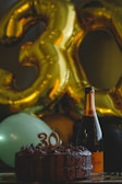 A festive setting features large golden balloons shaped like the number 30, accompanied by a chocolate cake with candles indicating the number 30. Beside the cake, there is a bottle of champagne. The ambiance suggests a celebration, likely a 30th birthday.