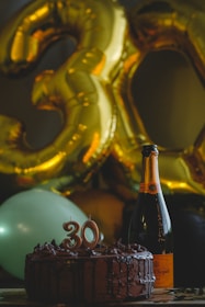 A festive setting features large golden balloons shaped like the number 30, accompanied by a chocolate cake with candles indicating the number 30. Beside the cake, there is a bottle of champagne. The ambiance suggests a celebration, likely a 30th birthday.