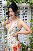 A woman stands confidently against a decorative lattice and foliage background, wearing a colorful floral dress. Her dark hair is styled in a loose, elegant updo, and she has dramatic earrings. Her expression is poised, with a hand resting near her collarbone.