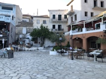 A charming Mediterranean street scene with outdoor dining tables set up on a stone-paved area. Surrounding the area are quaint, multi-story buildings with balconies and vibrant facades. Potted plants and trees add greenery to the scene, as several people sit at the tables, enjoying their time.