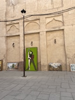 A collection of paintings placed against a textured, beige wall. The largest painting depicts a figure in green and brown standing in a dynamic pose. Additional paintings with landscapes and buildings are arranged beside it on a paved surface. A tall lamppost extends upwards, dividing the scene.