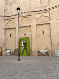 A collection of paintings placed against a textured, beige wall. The largest painting depicts a figure in green and brown standing in a dynamic pose. Additional paintings with landscapes and buildings are arranged beside it on a paved surface. A tall lamppost extends upwards, dividing the scene.