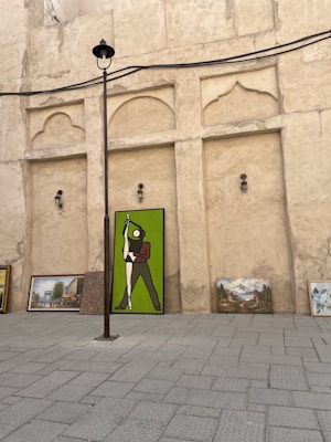 A collection of paintings placed against a textured, beige wall. The largest painting depicts a figure in green and brown standing in a dynamic pose. Additional paintings with landscapes and buildings are arranged beside it on a paved surface. A tall lamppost extends upwards, dividing the scene.