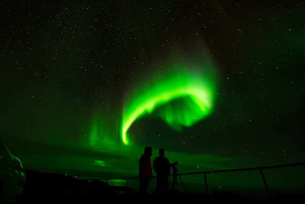 Two people with a tripod silhouetted against a bright green aurora curling over Nordkapp, Norway