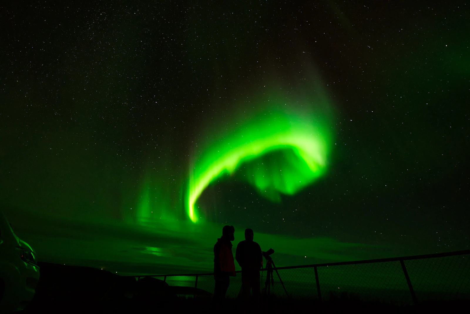 Two people with a tripod silhouetted against a bright green aurora curling over Nordkapp, Norway