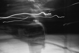 Blurred black and white image featuring streaks of light and shadow, creating an abstract and ethereal effect. The scene appears to contain motion with no clear focus, enhancing the mysterious atmosphere.