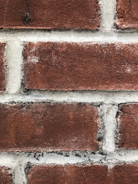Close-up view of a red brick wall with gray mortar lines. The texture of the brick is rough and weathered, showing signs of wear with patches of darker red and slight white remnants. The mortar between the bricks appears uneven and slightly discolored.