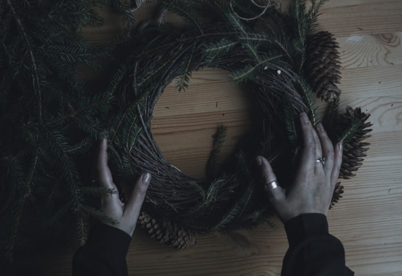 Pine branches and cones are being arranged into a circular wreath against a wooden surface. Hands are gently positioning the greenery, adding a natural and rustic touch to the scene.