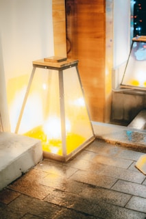 A large, modern lantern sits on a tiled floor, emitting a warm yellow glow that illuminates the surrounding area. The lantern is encased in a transparent material with a wooden top and metal frame. The background includes wooden panels and a soft light ambiance, suggesting a cozy indoor or semi-outdoor setting.