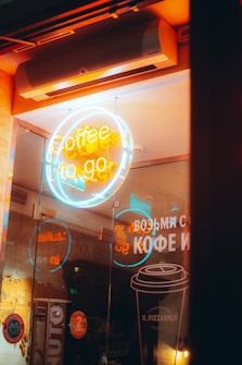 A brightly lit neon sign reading 'coffee to go' is displayed inside a glass storefront, surrounded by reflections and signage in a cozy coffee shop. The warm hues of orange contrast with the cool blue tones from the neon lights. Various decorative elements and text in another language are visible on the glass, contributing to an urban, inviting atmosphere.