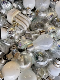 A collection of various types of light bulbs piled together, including incandescent, CFL, and LED bulbs. The bulbs are mostly clear or white, with some having metallic bases. The arrangement of the bulbs appears haphazard, showcasing different shapes and sizes.
