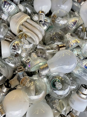 A collection of various types of light bulbs piled together, including incandescent, CFL, and LED bulbs. The bulbs are mostly clear or white, with some having metallic bases. The arrangement of the bulbs appears haphazard, showcasing different shapes and sizes.
