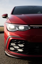 A close-up view of the front part of a red sports car, focusing on the headlights and grille. The headlights are modern and sharply designed, accompanied by a black, hexagonal-patterned grille. The car's badge is visible near the headlights, and the overall appearance is sleek and aggressive.