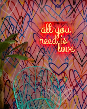 A vibrant wall with a multitude of overlapping heart shapes in various colors serves as the backdrop. In front of this, a neon sign with the phrase 'all you need is love' glows warmly in red. A green wire chair and a green plant with broad leaves are positioned nearby, adding an organic touch to the energetic scene.