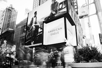 A bustling urban scene with a large American Eagle billboard prominently displayed on a building. The busy street is filled with blurred figures walking by, indicating motion and activity. Nearby, a vendor stand displays a variety of New York-themed merchandise including hats and shirts. The architecture features a mix of tall buildings, modern glass facades, and advertisements, showcasing a lively metropolitan environment.