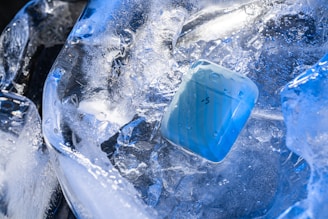 A small blue rectangular object, possibly a case, is partially submerged in a mass of chipped and solid ice. The ice exhibits a transparent and frosty appearance, capturing light reflections and creating a shimmering effect. The setting appears cold and crisp, with the ice dominating the scene.