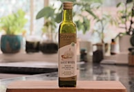 A bottle of Cobbam Estate garlic infused Australian extra virgin olive oil is placed on a wooden cutting board in a kitchen setting. The background features blurred greenery and kitchen items, creating a cozy and natural atmosphere.