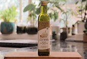 A bottle of Cobbam Estate garlic infused Australian extra virgin olive oil is placed on a wooden cutting board in a kitchen setting. The background features blurred greenery and kitchen items, creating a cozy and natural atmosphere.