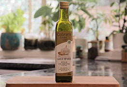 A bottle of Cobbam Estate garlic infused Australian extra virgin olive oil is placed on a wooden cutting board in a kitchen setting. The background features blurred greenery and kitchen items, creating a cozy and natural atmosphere.