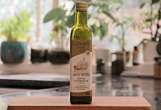 A bottle of Cobbam Estate garlic infused Australian extra virgin olive oil is placed on a wooden cutting board in a kitchen setting. The background features blurred greenery and kitchen items, creating a cozy and natural atmosphere.