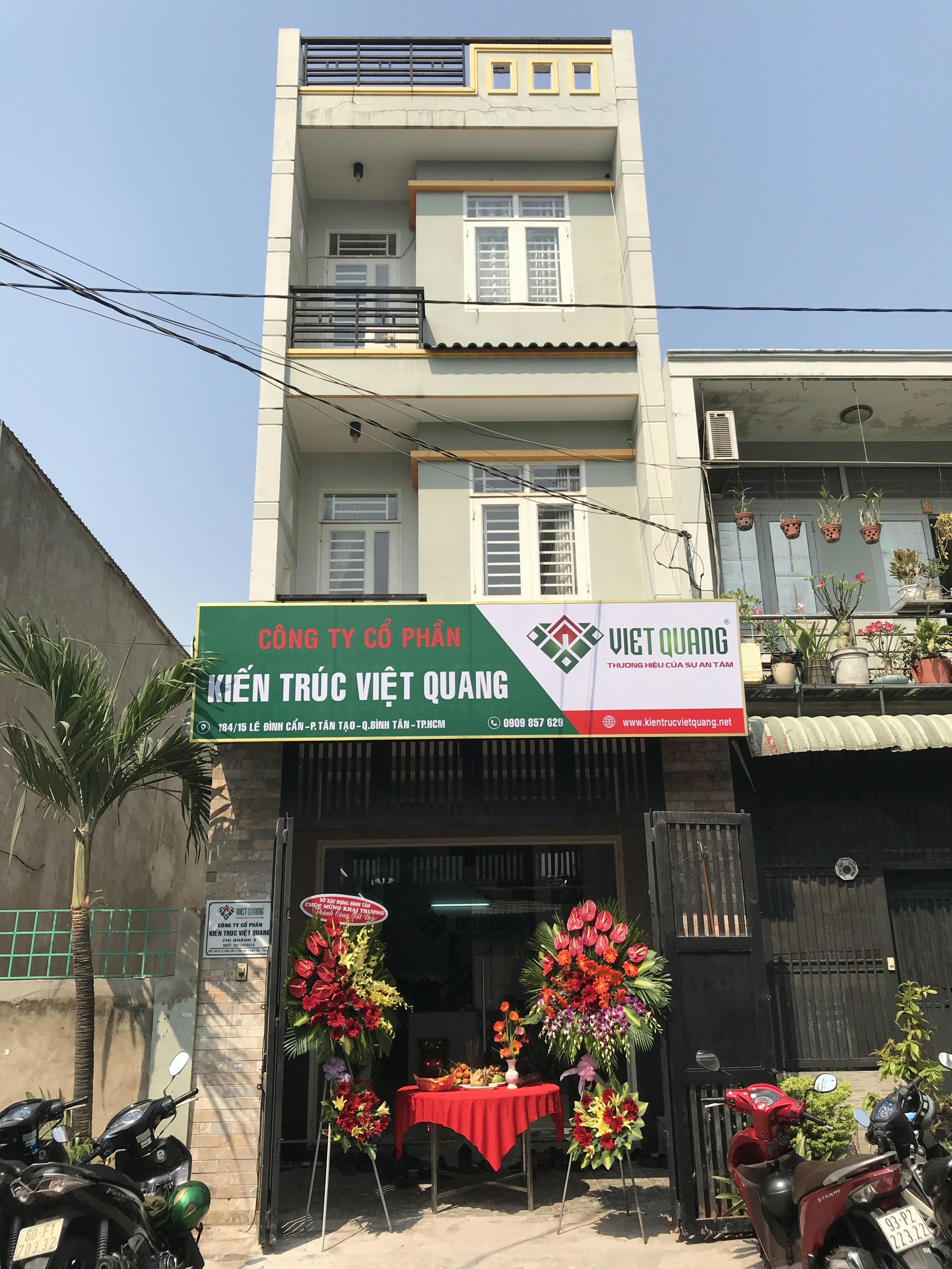 A multi-story building with a business on the ground floor displaying a sign for an architectural company. The facade includes balconies with railings. In front of the entrance, there is a table covered with a red cloth, adorned with decorative floral arrangements. Motorbikes are parked along the sidewalk and there are some potted plants on a ledge above the entrance.
