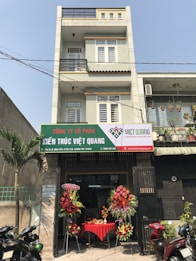 A multi-story building with a business on the ground floor displaying a sign for an architectural company. The facade includes balconies with railings. In front of the entrance, there is a table covered with a red cloth, adorned with decorative floral arrangements. Motorbikes are parked along the sidewalk and there are some potted plants on a ledge above the entrance.