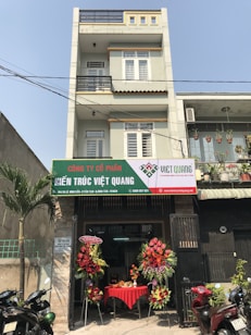 A multi-story building with a business on the ground floor displaying a sign for an architectural company. The facade includes balconies with railings. In front of the entrance, there is a table covered with a red cloth, adorned with decorative floral arrangements. Motorbikes are parked along the sidewalk and there are some potted plants on a ledge above the entrance.