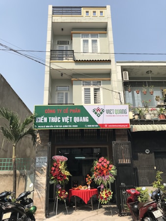 A multi-story building with a business on the ground floor displaying a sign for an architectural company. The facade includes balconies with railings. In front of the entrance, there is a table covered with a red cloth, adorned with decorative floral arrangements. Motorbikes are parked along the sidewalk and there are some potted plants on a ledge above the entrance.