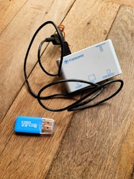 A white Transcend card reader with multiple slots and a black cable is placed on a wooden surface. Next to it is a blue Micro SD to USB 2.0 adapter.