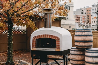 A wood-fired pizza oven with a chimney is situated outdoors. Nearby, several large barrels are stacked. The ground is covered in autumn leaves, and a tree with orange foliage stands beside the oven. String lights are wrapped around the tree trunk, and a wooden fence encloses the area. In the background, urban buildings are visible, suggesting the location is a city or town.