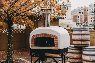 A wood-fired pizza oven with a chimney is situated outdoors. Nearby, several large barrels are stacked. The ground is covered in autumn leaves, and a tree with orange foliage stands beside the oven. String lights are wrapped around the tree trunk, and a wooden fence encloses the area. In the background, urban buildings are visible, suggesting the location is a city or town.