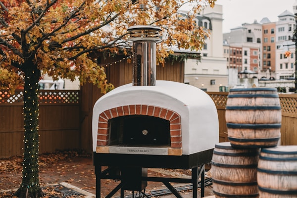 A wood-fired pizza oven with a chimney is situated outdoors. Nearby, several large barrels are stacked. The ground is covered in autumn leaves, and a tree with orange foliage stands beside the oven. String lights are wrapped around the tree trunk, and a wooden fence encloses the area. In the background, urban buildings are visible, suggesting the location is a city or town.