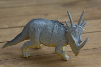 A plastic toy dinosaur with a textured surface rests on a wooden floor. The dinosaur has a gray body with prominent horns and a frilled head, resembling a ceratopsid.
