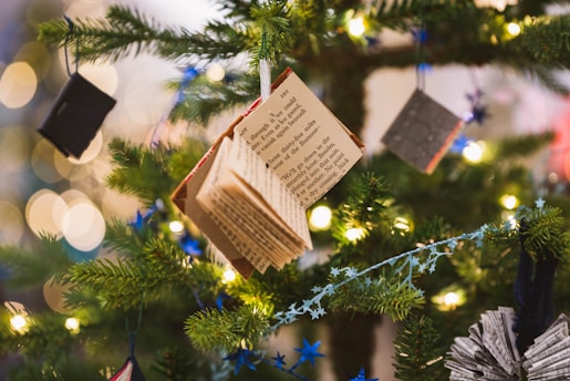 A close-up view of a Christmas tree adorned with unique decorations such as books hanging from the branches. The tree is lit with warm white fairy lights, and there are blue star garlands also draped around the branches. The background features soft bokeh circles from additional lighting, creating a festive and cozy atmosphere.