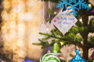 A festive setting features a decorated Christmas tree with ornaments, including a blue snowflake and green bauble. A handwritten note with the message 'you love to take care of others' is attached to the tree. The background is filled with warm, yellow bokeh lights, creating a cozy and cheerful atmosphere.