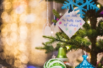A festive setting features a decorated Christmas tree with ornaments, including a blue snowflake and green bauble. A handwritten note with the message 'you love to take care of others' is attached to the tree. The background is filled with warm, yellow bokeh lights, creating a cozy and cheerful atmosphere.