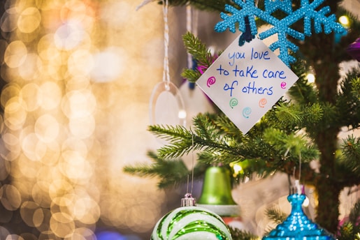 A festive setting features a decorated Christmas tree with ornaments, including a blue snowflake and green bauble. A handwritten note with the message 'you love to take care of others' is attached to the tree. The background is filled with warm, yellow bokeh lights, creating a cozy and cheerful atmosphere.