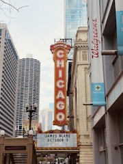 A city scene featuring a vintage-style marquee sign for an event on October 3rd, with tall buildings and a Walgreens pharmacy sign visible. The iconic Chicago Theatre sign is prominent, offering a sense of urban culture.