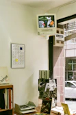 A small interior space featuring a CRT television with built-in VCR mounted high on the wall displaying an image of colorful flowers or abstract shapes. Below, a wood shelf holds various books and magazines, including one with 'bauhaus' text visible. A framed geometric artwork with black, yellow, and blue squares hangs on the wall. The window next to the shelf shows a partial view of the street outside with a passing car.