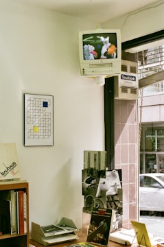 A small interior space featuring a CRT television with built-in VCR mounted high on the wall displaying an image of colorful flowers or abstract shapes. Below, a wood shelf holds various books and magazines, including one with 'bauhaus' text visible. A framed geometric artwork with black, yellow, and blue squares hangs on the wall. The window next to the shelf shows a partial view of the street outside with a passing car.