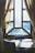 A wooden table and chairs are positioned in front of a large, finely crafted window with multiple panes, framed by elegant, draped curtains. Sunlight streams in, casting soft, diffused shadows on the table and creating an inviting and warm atmosphere.