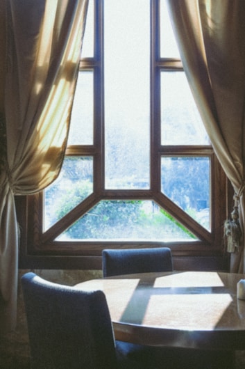 A wooden table and chairs are positioned in front of a large, finely crafted window with multiple panes, framed by elegant, draped curtains. Sunlight streams in, casting soft, diffused shadows on the table and creating an inviting and warm atmosphere.