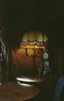 A dimly lit vintage setting with an ornate table lamp featuring a decorative patterned shade. Next to it, there is an intricately designed antique clock. The soft glow from the lamp highlights the textures and details of the objects, creating a cozy and nostalgic atmosphere. Rich fabrics are draped around the scene, enhancing the classical ambiance.